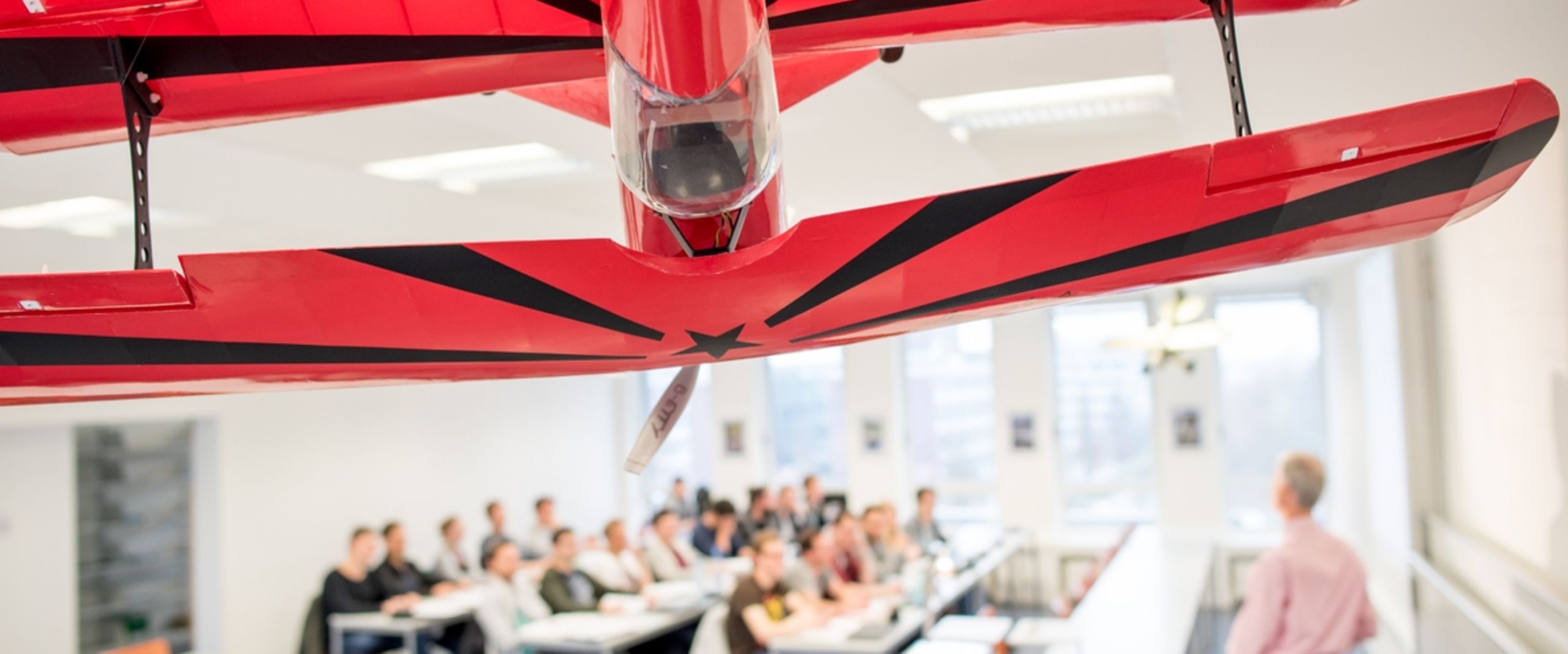 Auf dem Bild ist ein großes rotes Modellflugzeug zu sehen, das von der Decke eine Vorlesungssaales hängt. Im Hintergrund ist eine Grube Studierender und ein Lehrender im Seminarraum zu sehen.