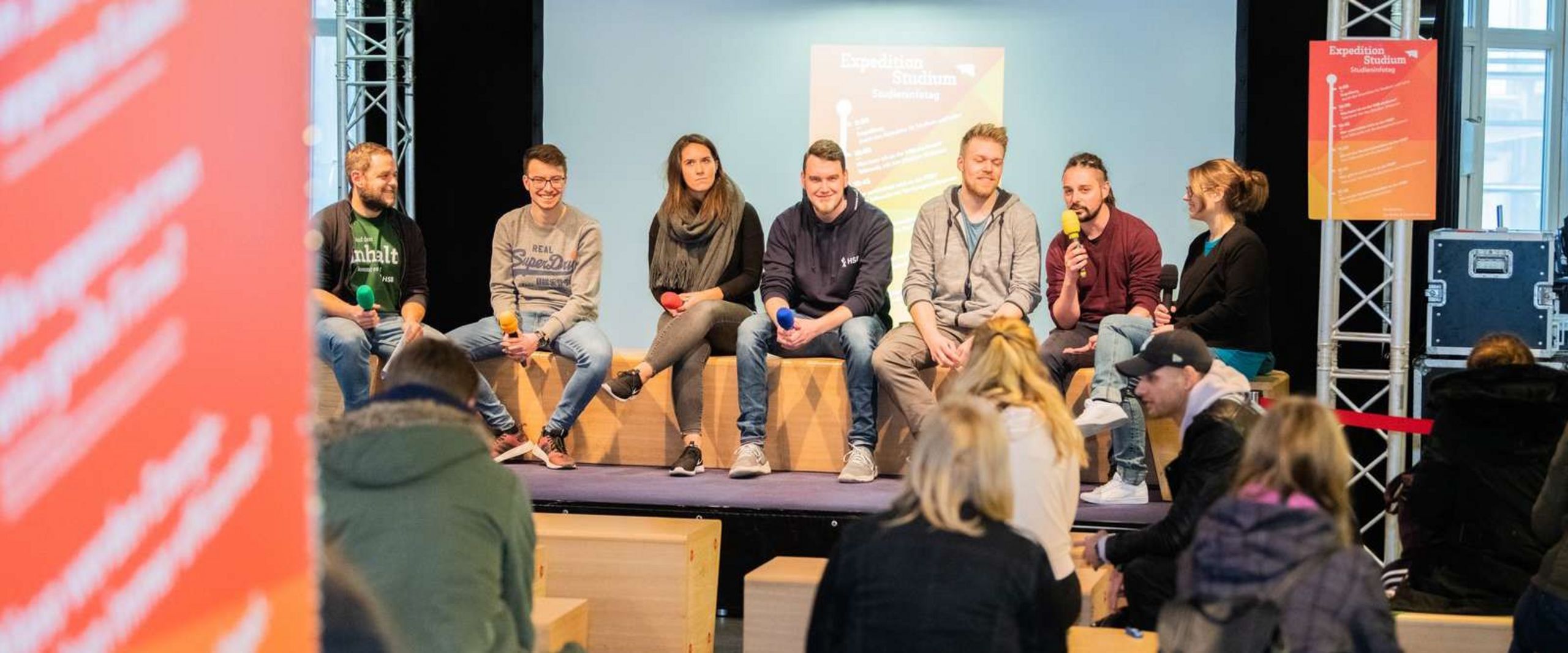 Sieben Studierende sitzen auf einer Bühne. Sie haben bunte Mikrofone in der Hand. Ein Student spricht in sein Mikrofon. Vor der Bühne sitzen einige Studieninteressierte, manche schauen zur Bühne, manche sprechen miteinander.