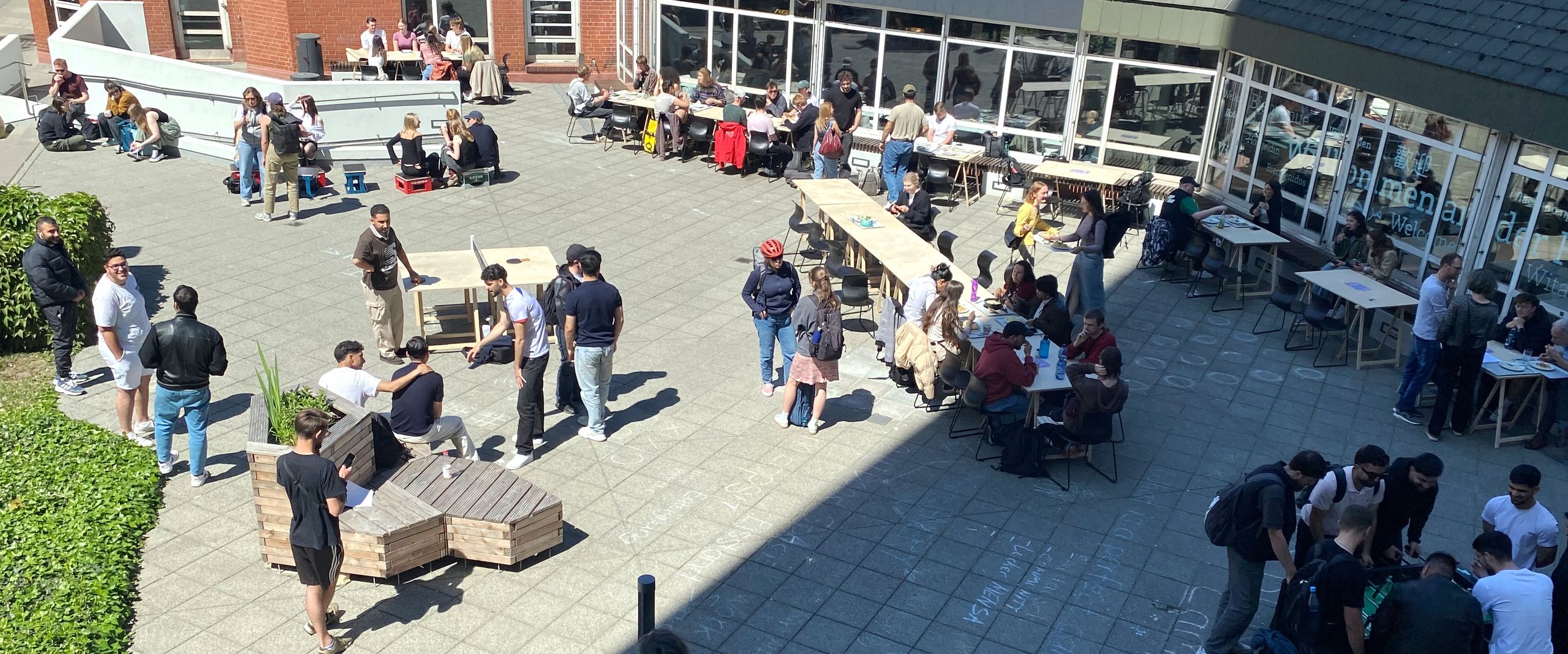 Photo from above on the forecourt of a building. People sit and stand there and play ping pong