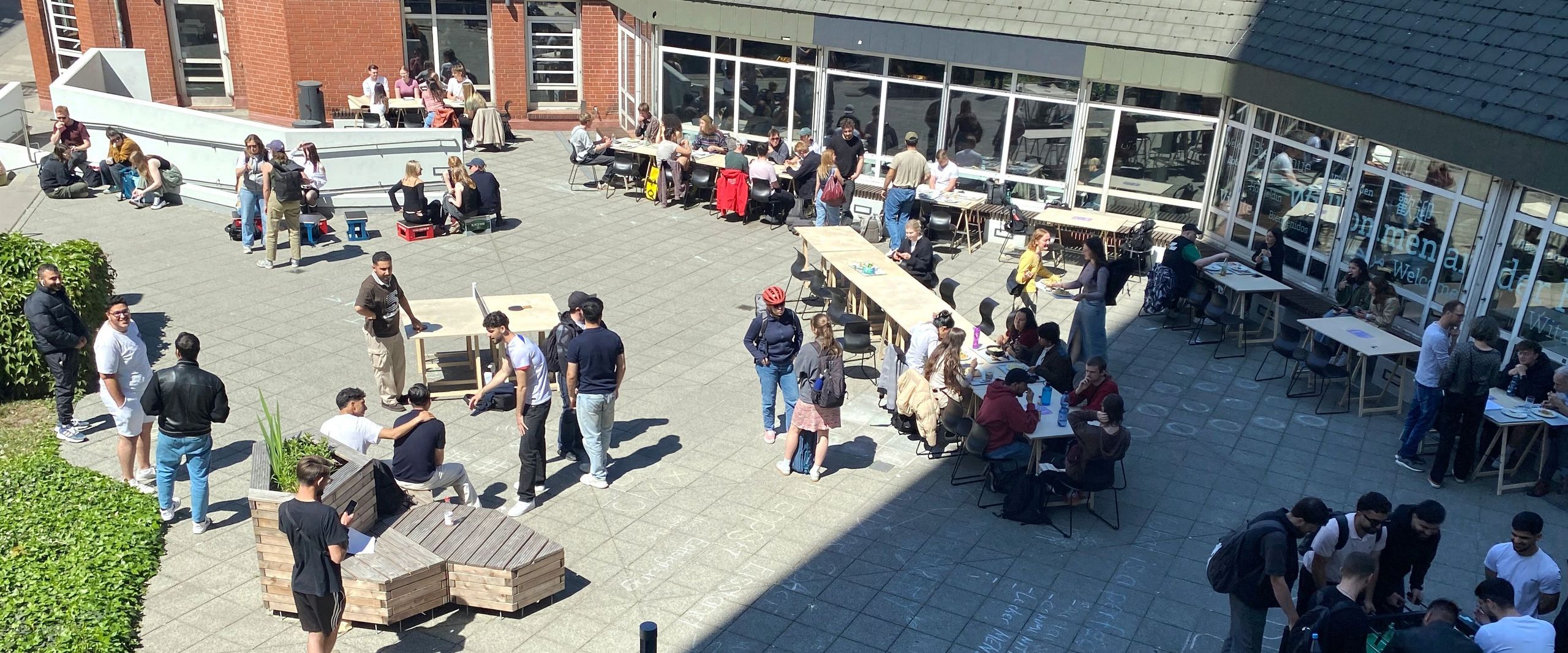 Bild von oben auf Vorplatz von Gebäude. Menschen sitzen und stehen dort und spielen Tischtennis