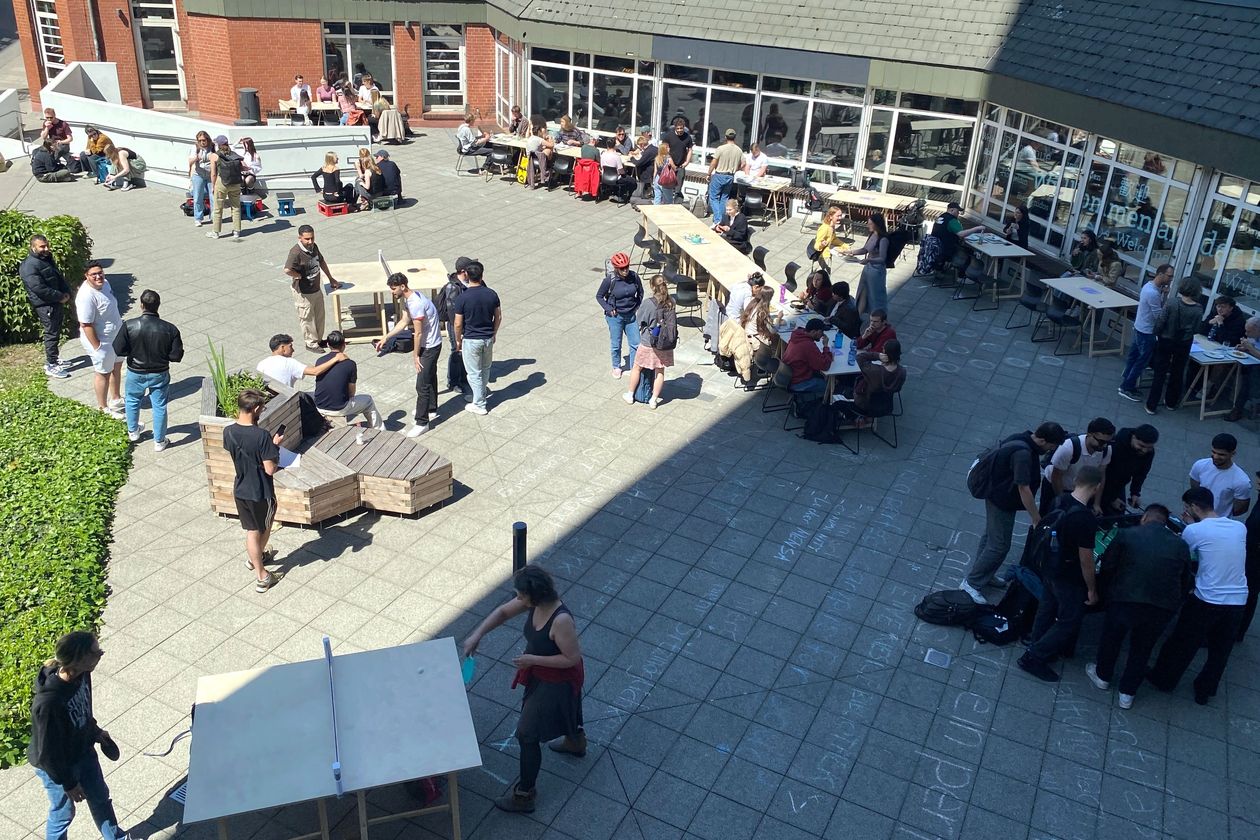 Photo from above on the forecourt of a building. People sit and stand there and play ping pong