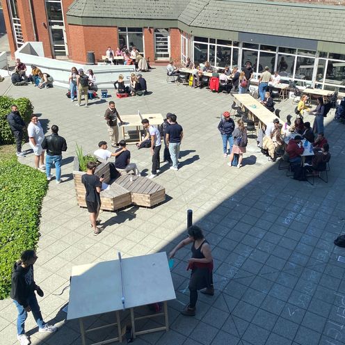 Photo from above on the forecourt of a building. People sit and stand there and play ping pong