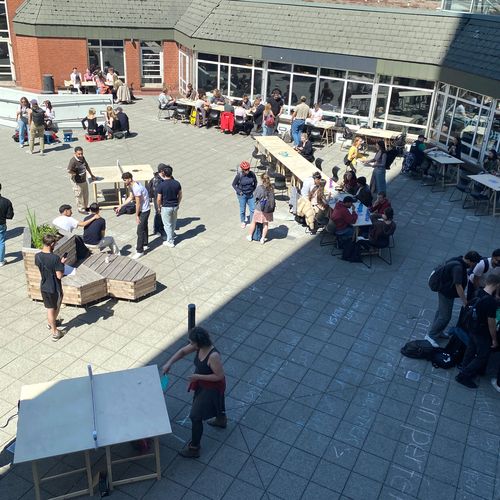 Bild von oben auf Vorplatz von Gebäude. Menschen sitzen und stehen dort und spielen Tischtennis