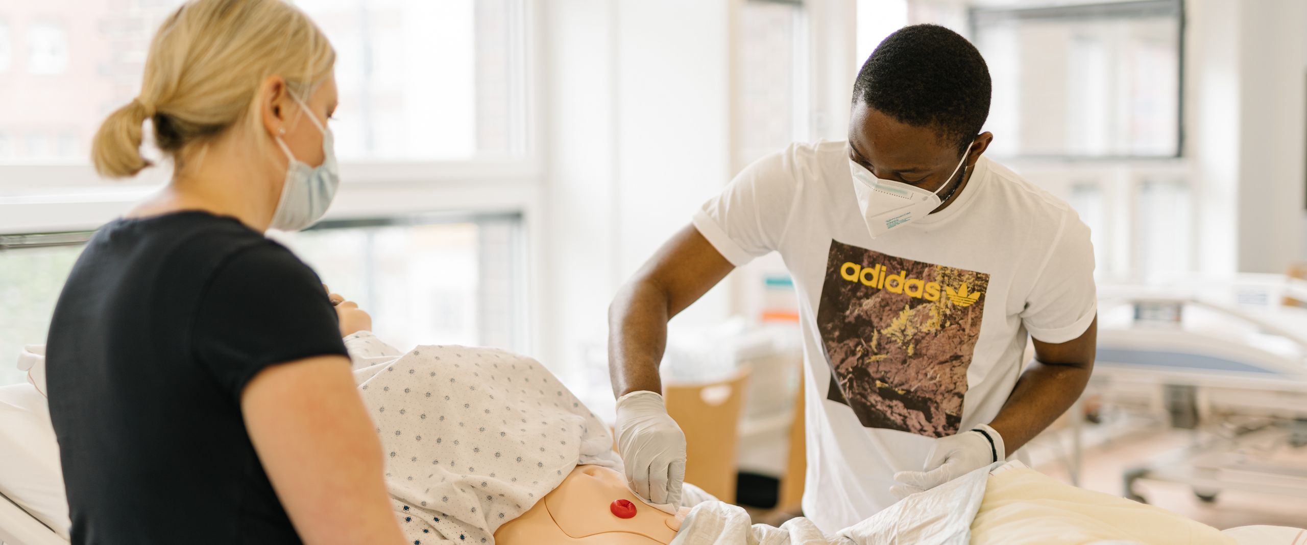 Ausbildungssituation am Pflegebett: Junge Frau und junger Mann &uuml;ben die Versorgung eines pflegbed&uuml;ftigen Patienten am Modell.