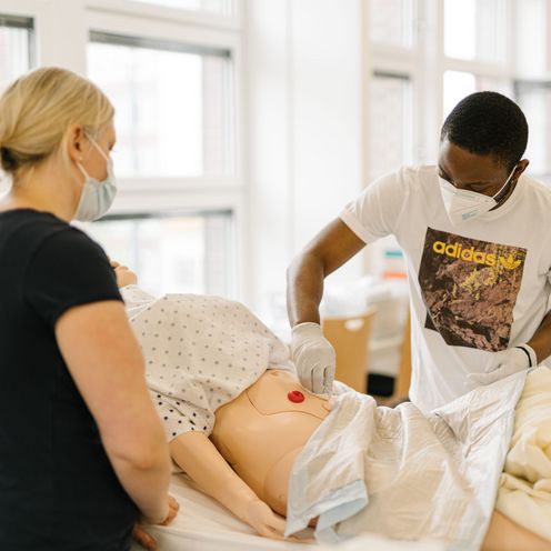 Ausbildungssituation am Pflegebett: Junge Frau und junger Mann &uuml;ben die Versorgung eines pflegbed&uuml;ftigen Patienten am Modell.