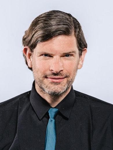 Auf dem Bild ist Björn Gering zusehen. Er hat braune Haare und trägt ein schwarzes Hemd mit einer blauen Krawatte. 