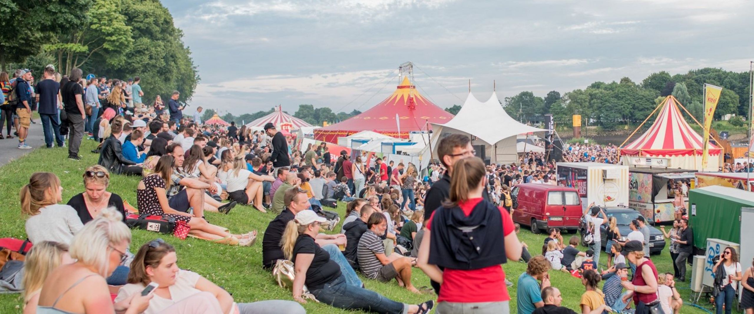 Menschen sitzen bei sommerlichen Temperaturen auf dem Deich. Im Hintergrund sieht man Festivalzelte.