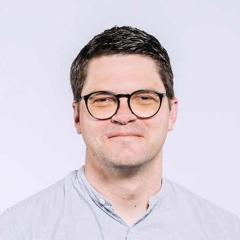 Auf dem Bild ist Christian Fieberg zu sehen. Er trägt kurzes schwarzes Haar, eine ründliche Brille und ein hell blaues Hemd. 