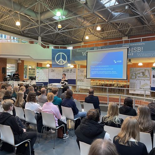 Saal mit zahlreichen G&auml;sten, die auf St&uuml;hlen sitzen. Vorne steht eine Frau und h&auml;lt eine Rede.