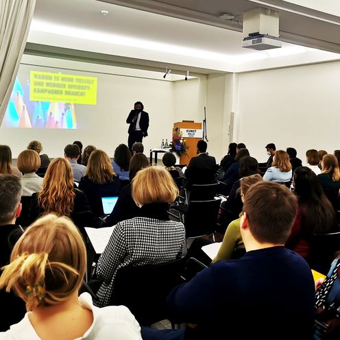 Menschen stehen in einem Foyer am STehtischen und reden. An den W&auml;nden h&auml;ngen Bilder