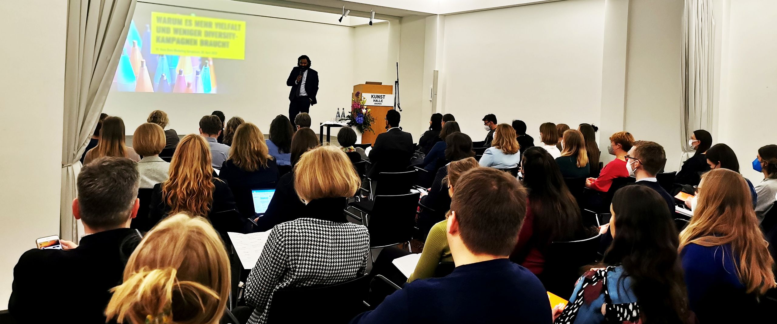 Menschen stehen in einem Foyer am STehtischen und reden. An den W&auml;nden h&auml;ngen Bilder