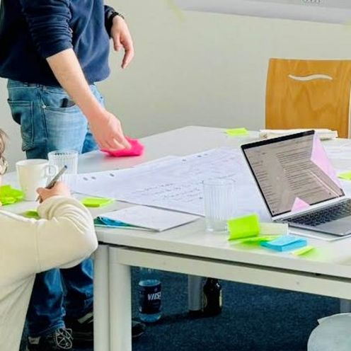 Junge Menschen im Stehen und Sitzen arbeiten an einem Tisch. An den W&auml;nden h&auml;ngen Plakate.