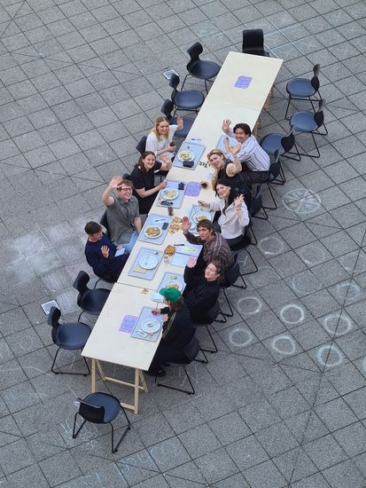 Bild aus der Vogelperspektive: Junge Männer und Frauen sitzen auf einem Platz an einerm langen Tisch mit Essenstabletts und winken hoch in die Kamera.
