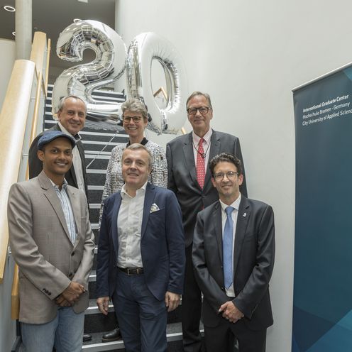 gruppenbild mit M&auml;nnern und Frauen auf einer Treppe. Hinter ihnen schwebt eine 20 als zwei silberne Luftballons.