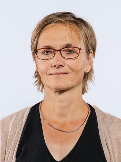 Auf dem Foto ist Regine Komoss zusehen. Sie hat kurzes blondes Haar und trägt eine Brille, sowie ein schwarzes T-Shirt unter einer hellbraunen Strickjacke. 