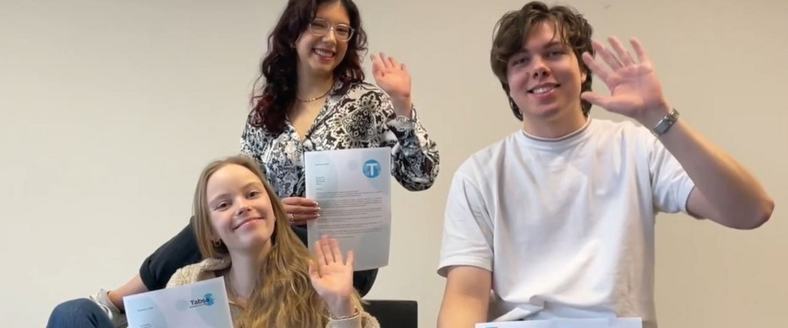 Zwei junge Frauen und ein junger Mann sitzen und winken. Sie halten in der Hand Urkunden mit Logos