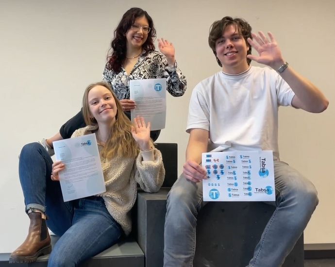 Zwei junge Frauen und ein junger Mann sitzen und winken. Sie halten in der Hand Urkunden mit Logos