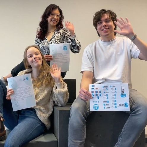 Zwei junge Frauen und ein junger Mann sitzen und winken. Sie halten in der Hand Urkunden mit Logos