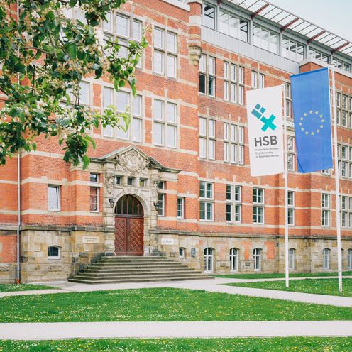 Rotes Backsteingeb&auml;ude an einer Wiese. Davor stehen Fahnen mit HSB-Logo und EU.