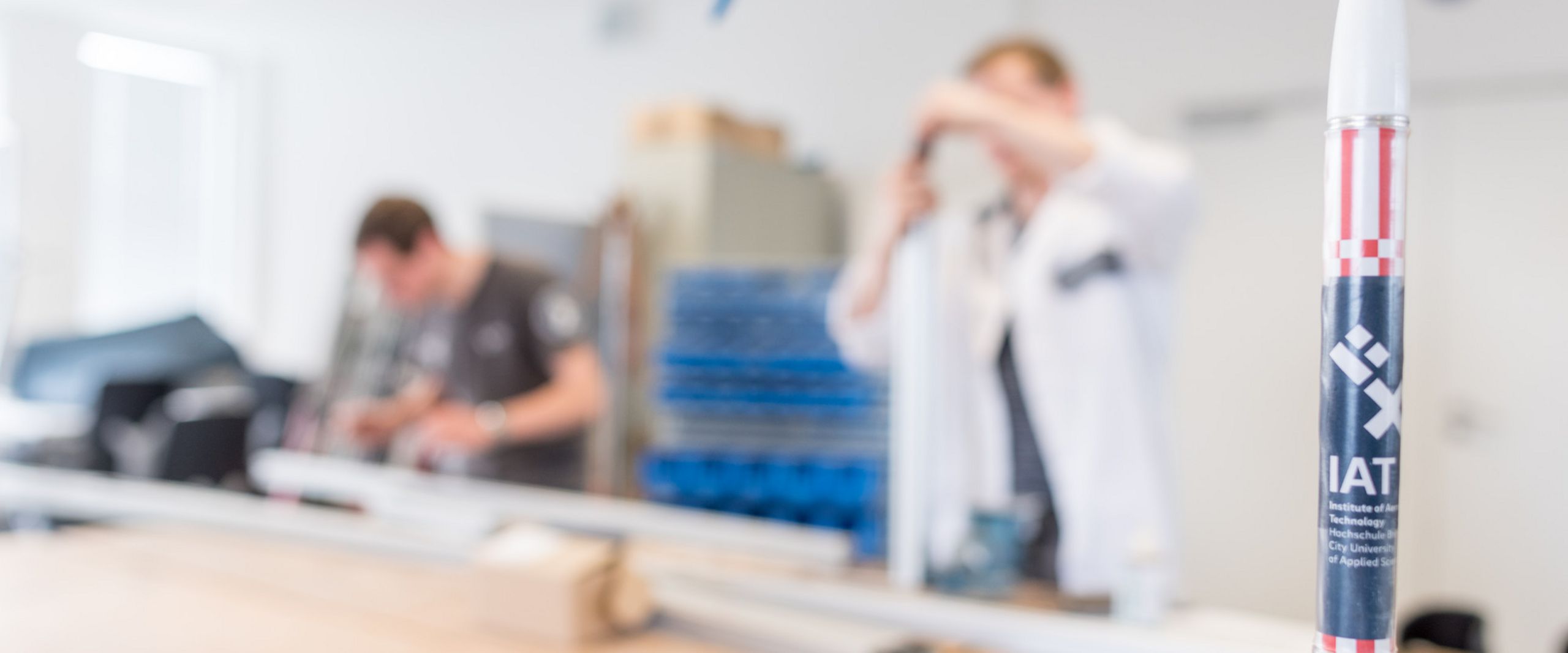 Studierende arbeiten an mechanischen Bauteilen in einer Werkstatt. Im Vordergrund steht das Miniaturmodell einer Rakete.