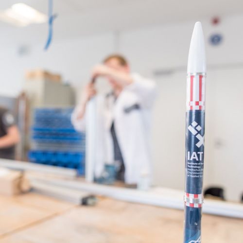 Studierende arbeiten an mechanischen Bauteilen in einer Werkstatt. Im Vordergrund steht das Miniaturmodell einer Rakete.