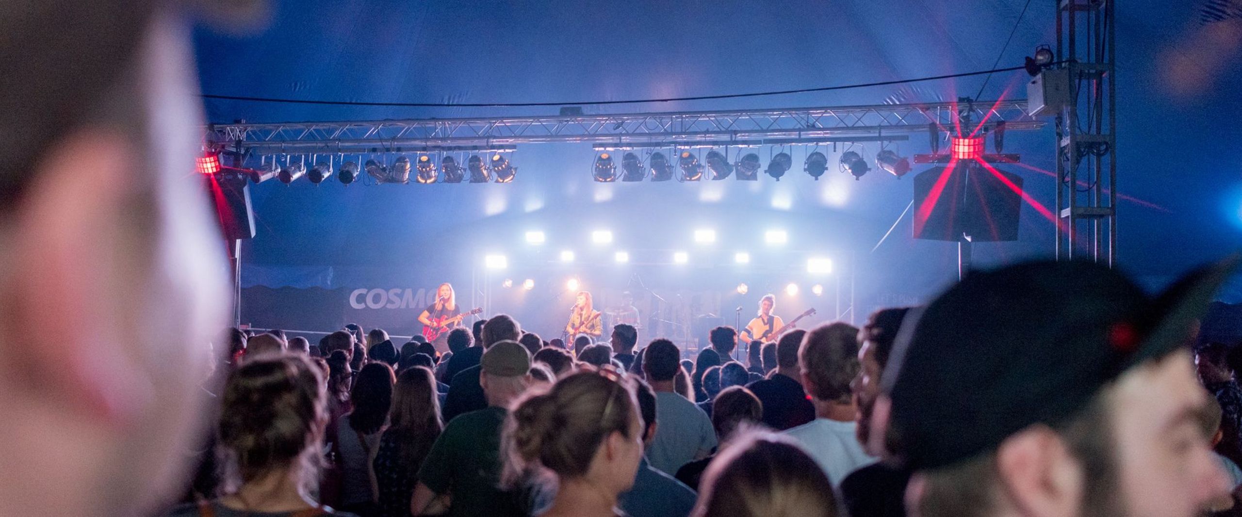 Ein großes Publikum steht vor einer Bühne in einem Zelt, auf der eine Band spielt.