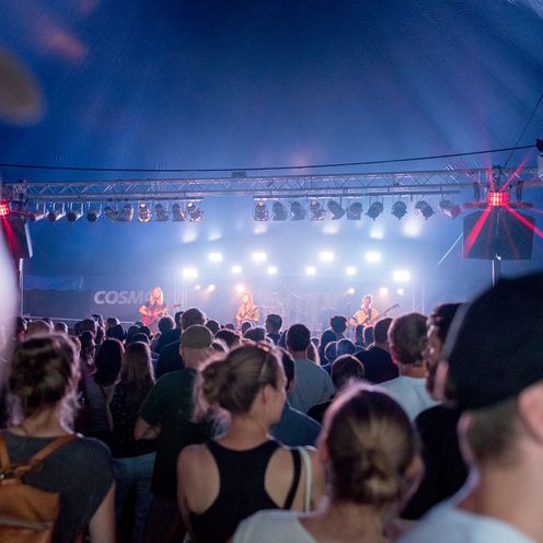 Ein gro&szlig;es Publikum steht vor einer B&uuml;hne in einem Zelt, auf der eine Band spielt.