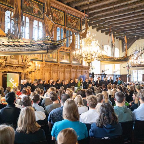 Überblick Erstsemesterbegrüßung 2024 Festassal mit vielen Gästen. Von der Decke hängen alte Holzschiffmodelle. Vorne steht ein Chor.