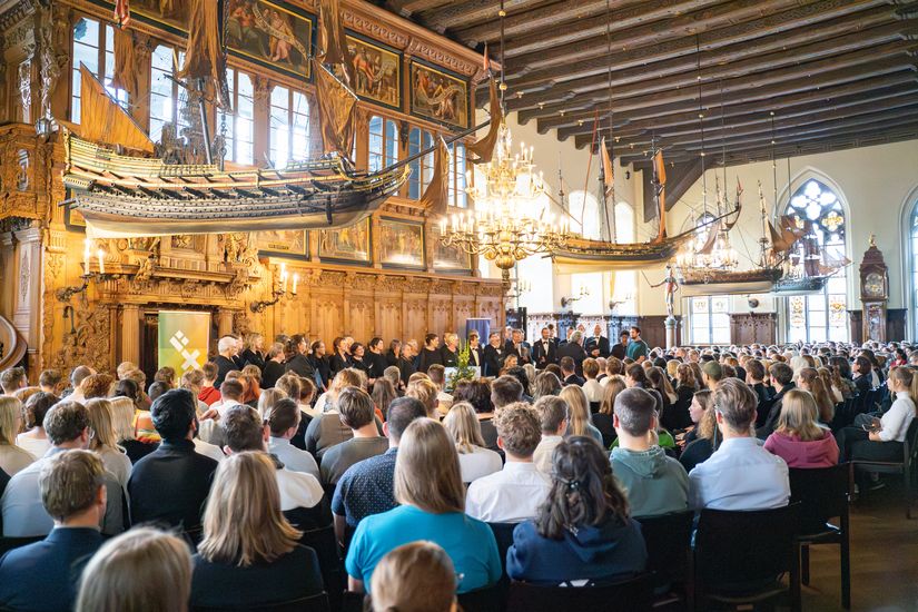 The picture shows the upper town hall, filled with HSB students and staff. Model ships hang from the ceiling. A choir stands in front 