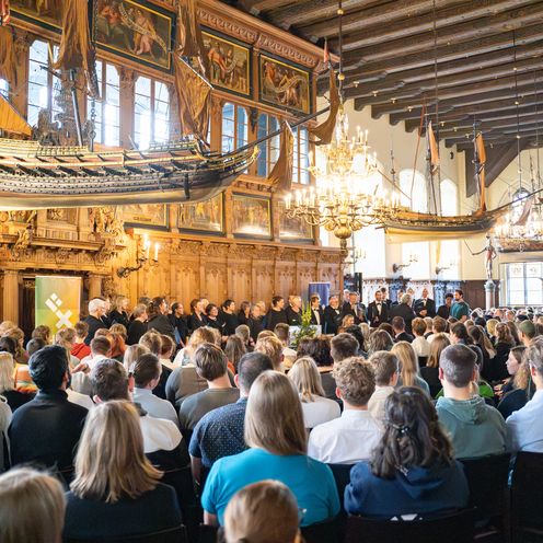 Festassal mit vielen G&auml;sten. Von der Decke h&auml;ngen alte Holzschiffmodelle. Vorne steht ein Chor.