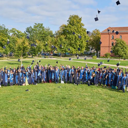 Auf dem Bild sind die Absolvent:innen des IGC zu sehen. Sie stehen auf einer gr&uuml;nen Wiese und Tragen alle Roben. Jubelt werfen sie ihre M&uuml;tzen in die Luft. 