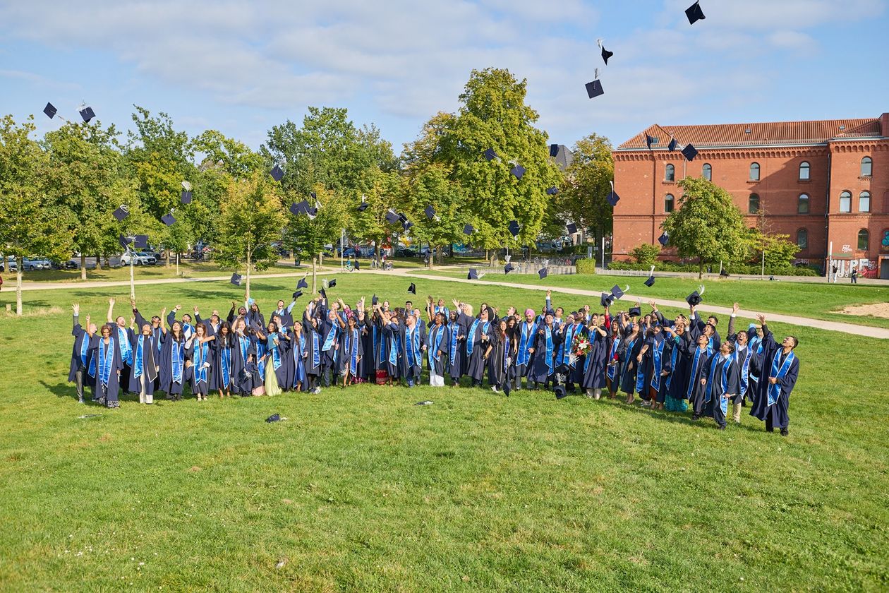 Auf dem Bild sind die Absolvent:innen des IGC zu sehen. Sie stehen auf einer grünen Wiese und Tragen alle Roben. Jubelt werfen sie ihre Mützen in die Luft. 