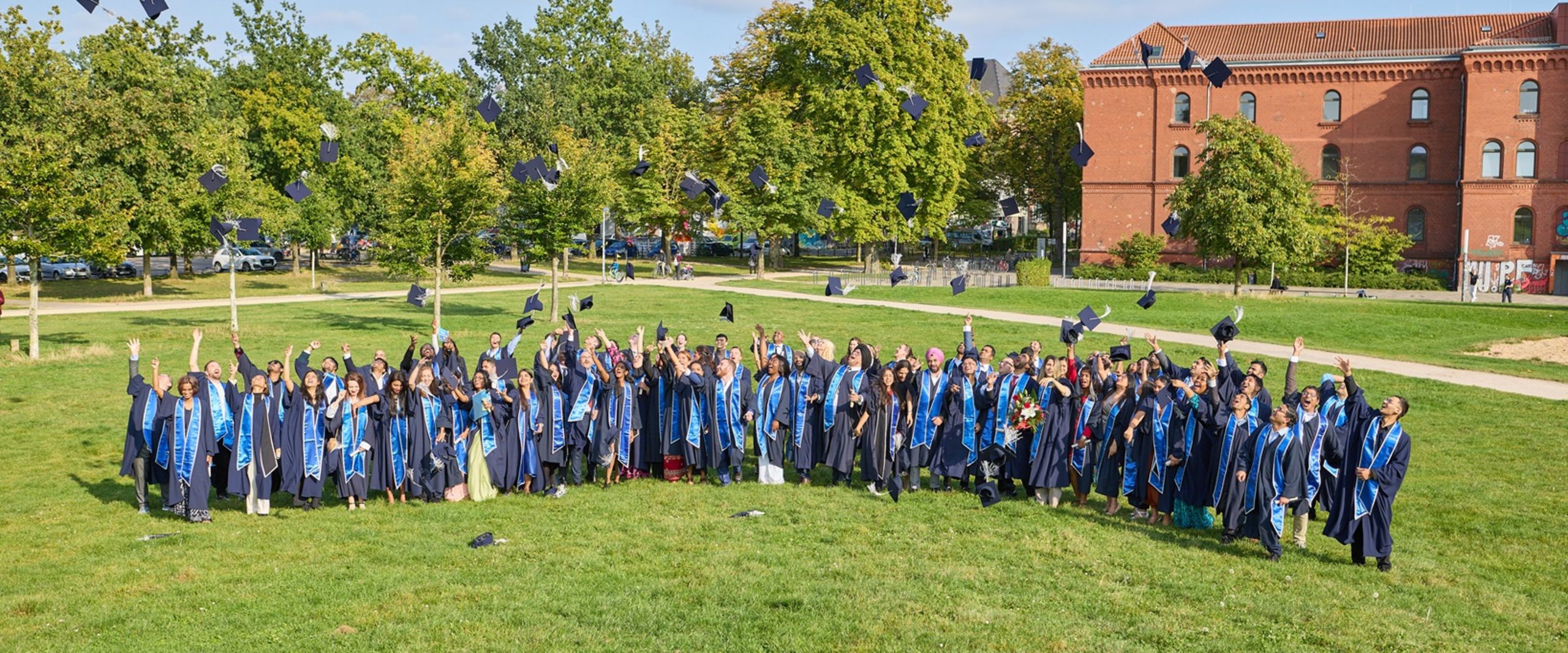 IGC Graduation 2024 Auf dem Bild sind die Absolvent:innen des IGC zu sehen. Sie stehen auf einer grünen Wiese und Tragen alle Roben. Jubelt werfen sie ihre Mützen in die Luft.