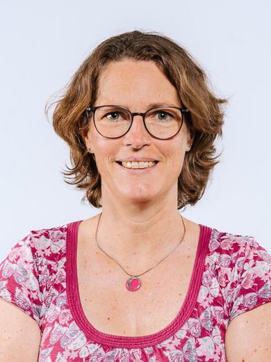 Auf dem Bild ist Meike Mossig zusehen. Sie hat kinnlange braune Haare. Sie trägt eine Brille und ein Rosa geblümtes Kleid mit einer passenden Kette. 