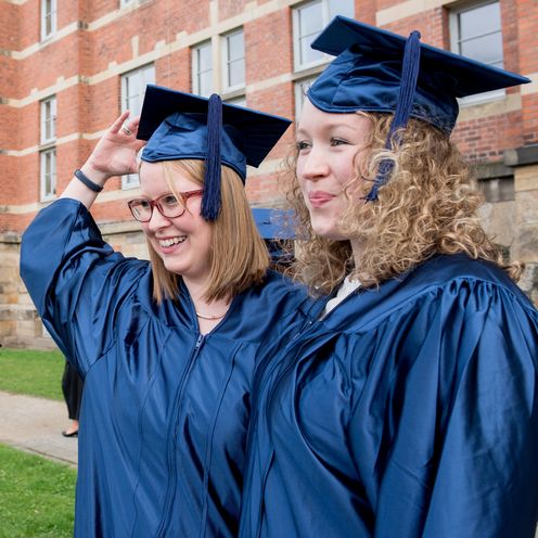 Zwei weibliche Studierende im Talar stehen vor dem M-Geb&auml;ude der HSB und posieren f&uuml;r ein Foto.