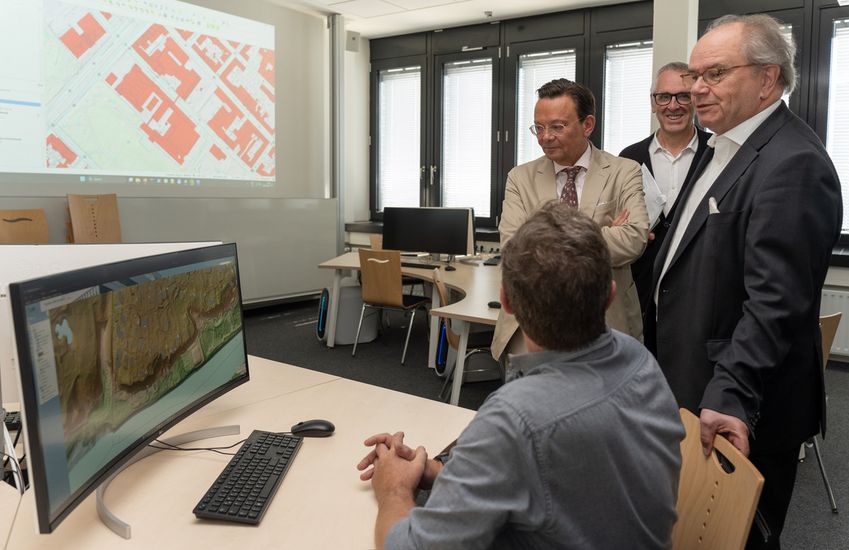 Drei Männer stehen an einem Schreibtisch. daran sitzt ein Mann an einem Bildschirm mit dem Rücken zur Kamera. An der Wand sieht man eine Grafik.