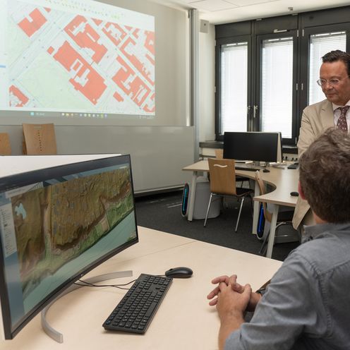 Drei M&auml;nner stehen an einem Schreibtisch. daran sitzt ein Mann an einem Bildschirm mit dem R&uuml;cken zur Kamera. An der Wand sieht man eine Grafik.