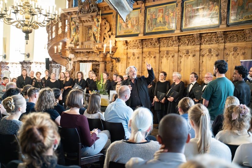 Chorleiter und Chor stehen vor Publikum in einem großen Saal