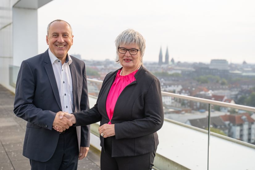 Man and woman shake hands and smile into the camera. They are standing on a roof terrace. In the background you can see the backdrop of the city centre of Bremen