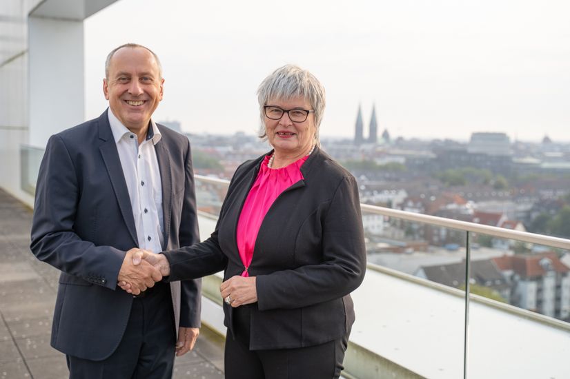 Mann und Frau geben sich die Hand und lächeln in die Kamera. Sie stehen auf einer Dachterrasse. Im Hintergrund sieht man die Kulisse der Innenstadt von Bremen