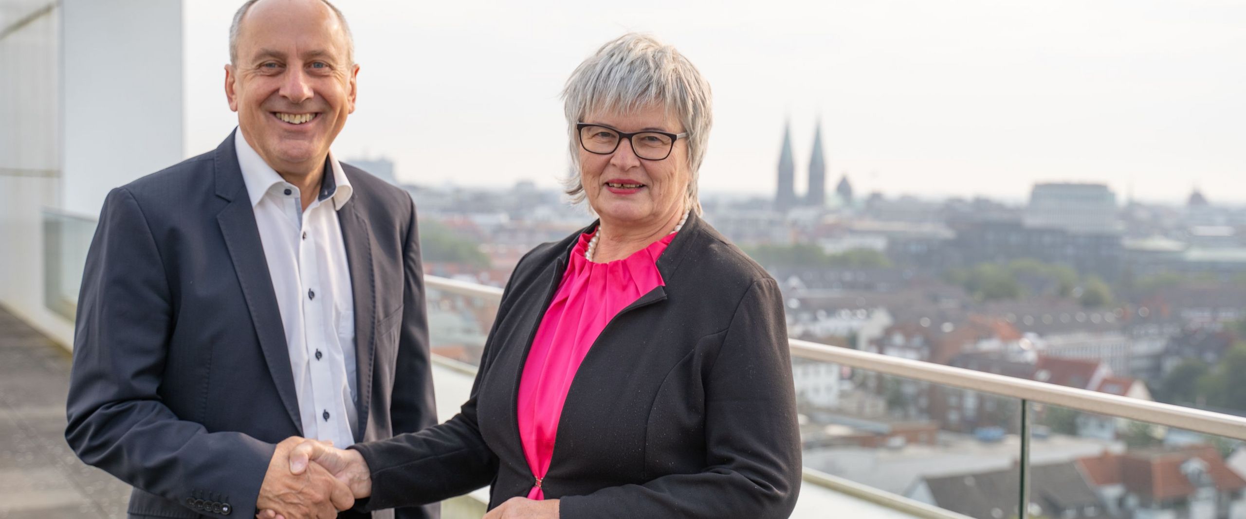 Mann und Frau geben sich die Hand und lächeln in die Kamera. Sie stehen auf einer Dachterrasse. Im Hintergrund sieht man die Kulisse der Innenstadt von Bremen