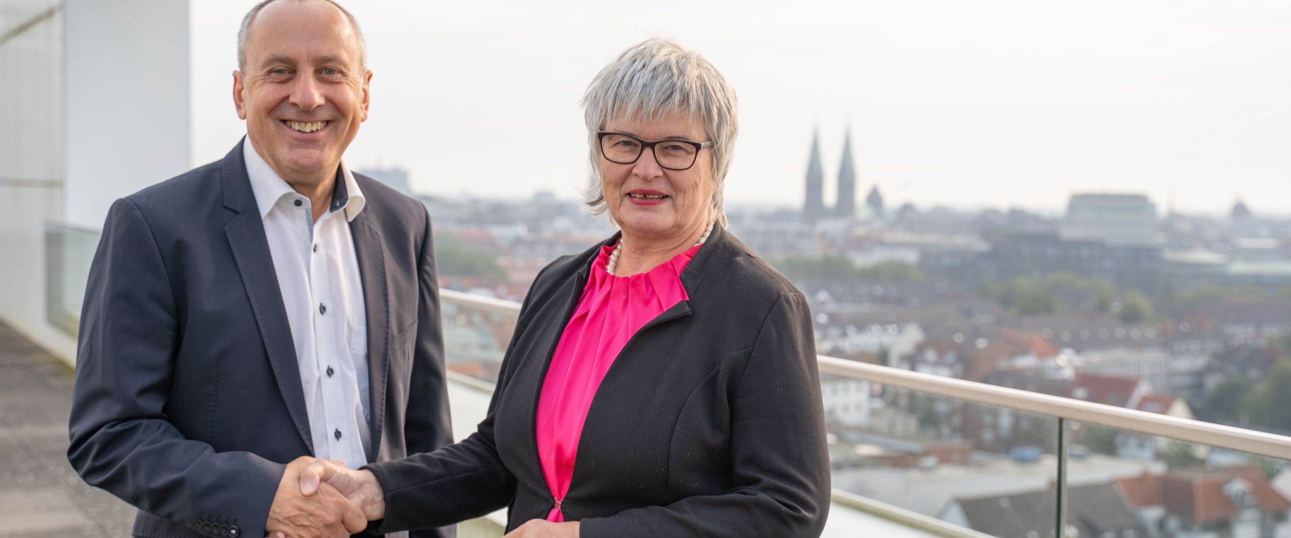 Man and woman shake hands and smile into the camera. They are standing on a roof terrace. In the background you can see the backdrop of the city centre of Bremen