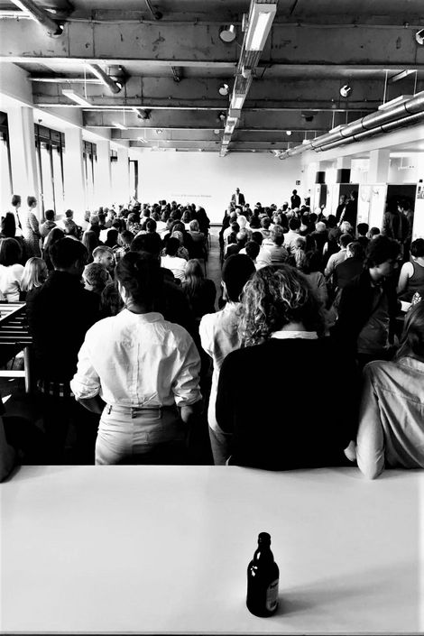 Ein Vortrag in einem Raum mit zahlreichen Personen. Im Vordergrund auf dem Tisch steht eine Flasche Bier.