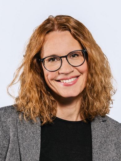 Auf dem Bild ist Ulrike Jacobs zusehen. Sie hat rötliches lockiges Haar, das bis zur Schulter reicht. Sie trägt eine Brille und einen grauen Blazer über einem schwarzen Shirt.