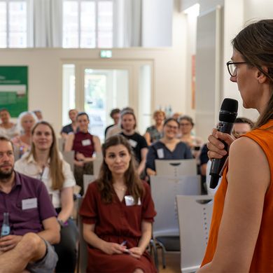Vortrag vor ca. 50 Workshop-Teilnehmenden in einem gro&szlig;en Raum. Im Vordergrund die Vortragende mit Mikrofon in der Hand.
