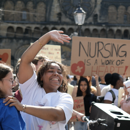 Auf dem Bild ist eine Demo auf dem Bremer Marktplatz zusehen. Im Vordergrund stehen zwei junge Frauen, eine von beiden grinst stark und winkt einer Person zu. Beide tragen Kasaks. Im Hintergrund sind Menschen und Pappschilder zusehen. 