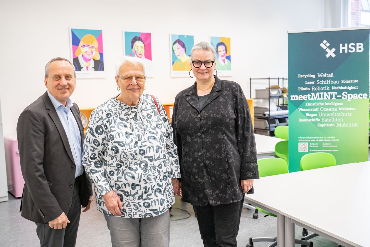 gruppenbild mit Mann und zwei Frauen. Im Hintergrund sieht man Tische und bunte Portraits von ber&uuml;hmten MINT-Wissenschaftlerinnen. rechts steht ein gr&uuml;n-wei&szlig;es Roll-up.