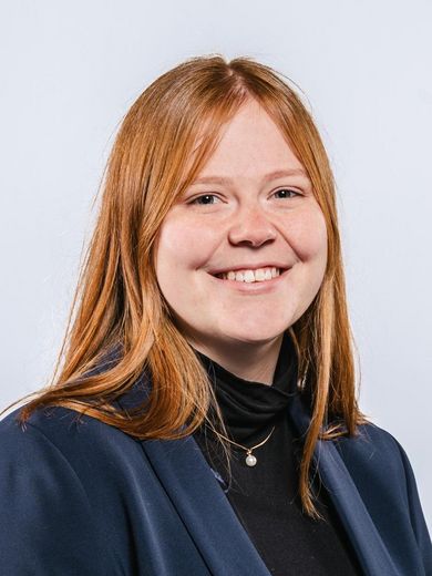 Auf dem Bild ist Michelle Mrosek zusehen. Sie hat lange rote Haare und trägt einen dunkelblauen Blazer über einem schwarzen Rollkragenpullover. 