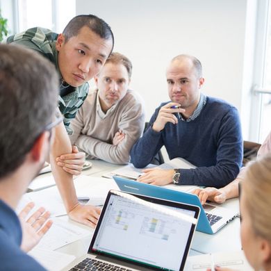 Mehrere Studenten sind um einen Tisch versammelt mit Unterlagen sowie Laptops und befinden sich in einer Diskussion.