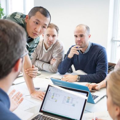 Mehrere Studenten sind um einen Tisch versammelt mit Unterlagen sowie Laptops und befinden sich in einer Diskussion.
